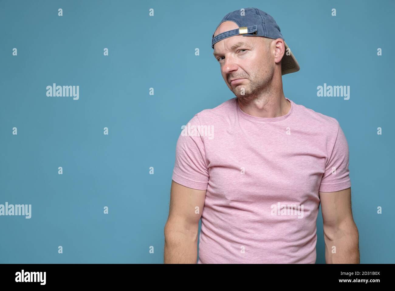 Suspicious Caucasian man in a cap and a pink T-shirt looks with disbelief. Copy space. Blue background. Stock Photo