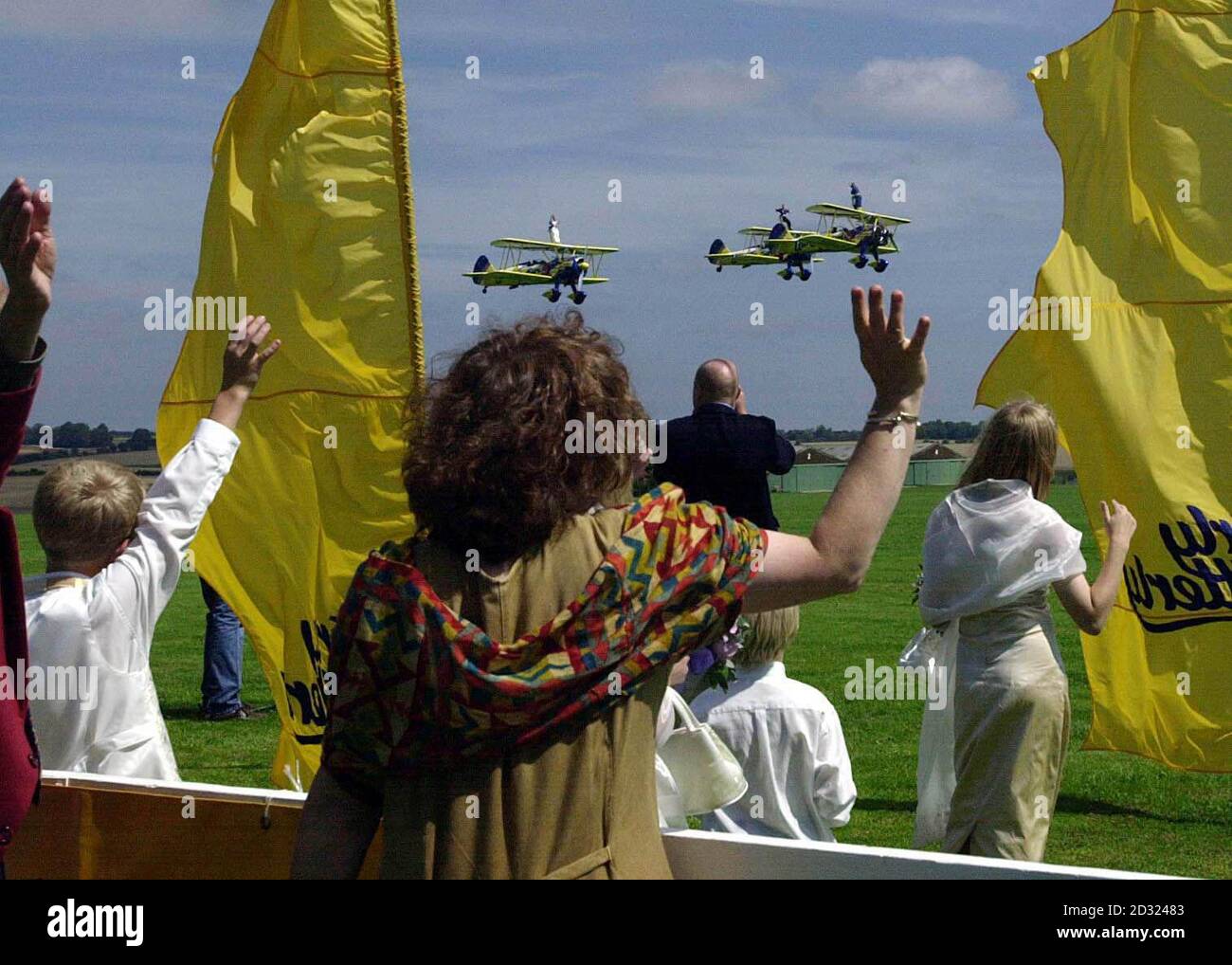 Family and friends wave at high-flying couple Caroline Hackwood (left plane) and Justin Bunn during their airborne wedding ceremony strapped to the wings of 1940s biplanes, with clergyman the Rev George Brigham on the front plane at Rendcomb airfield. * near Cirencester, Gloucestershire. Their their vows were relayed by microphones to their wedding party on the ground. Water board worker Caroline, 27, who was wearing a cream brocade and satin dress, screamed 'I do' as Mr Brigham shouted out the vows over loud speakers. The couple's 75 wedding guests on the ground clapped and cheered as the Stock Photo