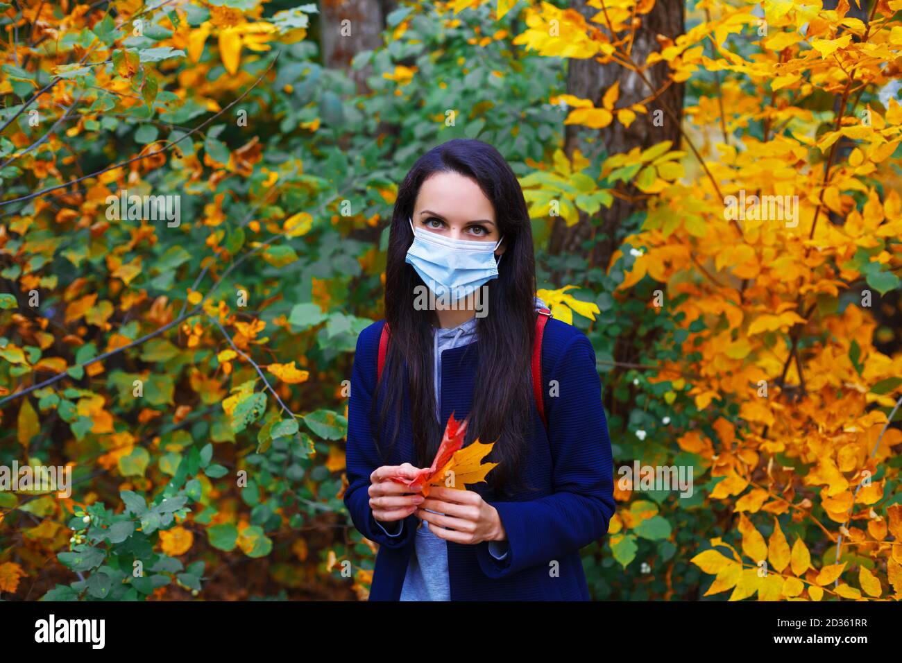Beautiful woman in protective mask holding maple leaves in colorful autumn forest. New normal concept. Stock Photo
