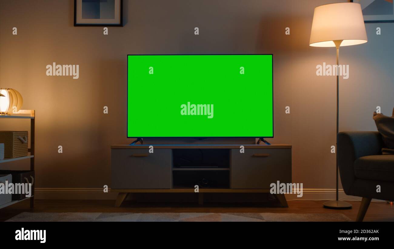 Shot of a TV with Horizontal Green Screen Mock Up. Cozy Evening Living Room with a Chair and Lamps Turned On at Home. Stock Photo