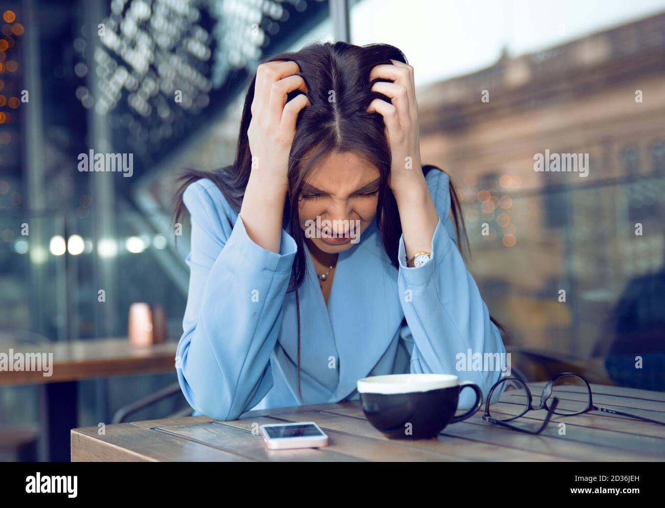 Stress. Portrait stressed sad young woman with coffee cup, phone and glasses sitting at table about to cry. City urban life style stress. Negative hum Stock Photo