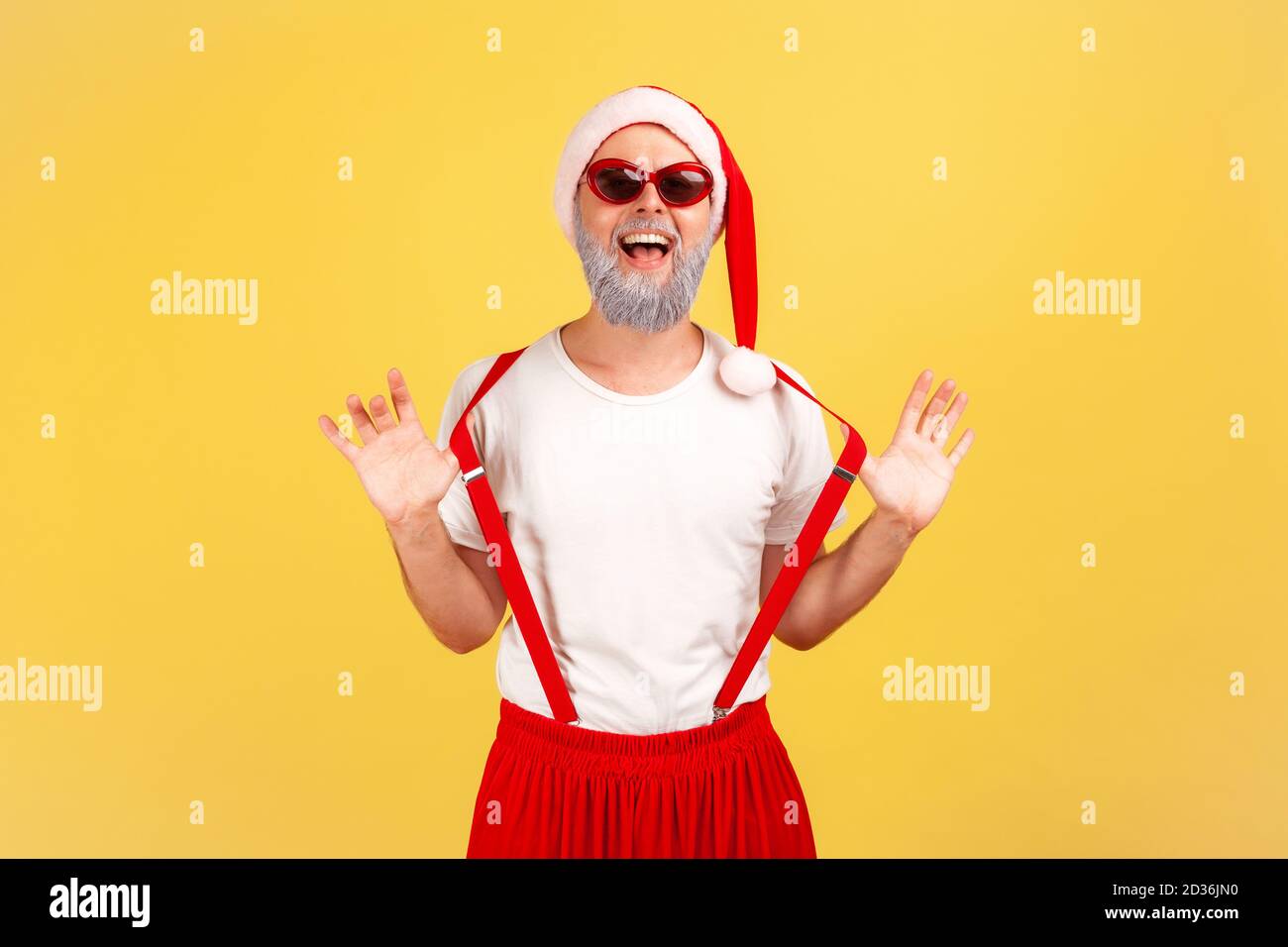 Funny cheerful elderly man in santa claus hat and stylish sunglasses holding his suspenders with hands and looking at camera with toothy smile. Indoor Stock Photo