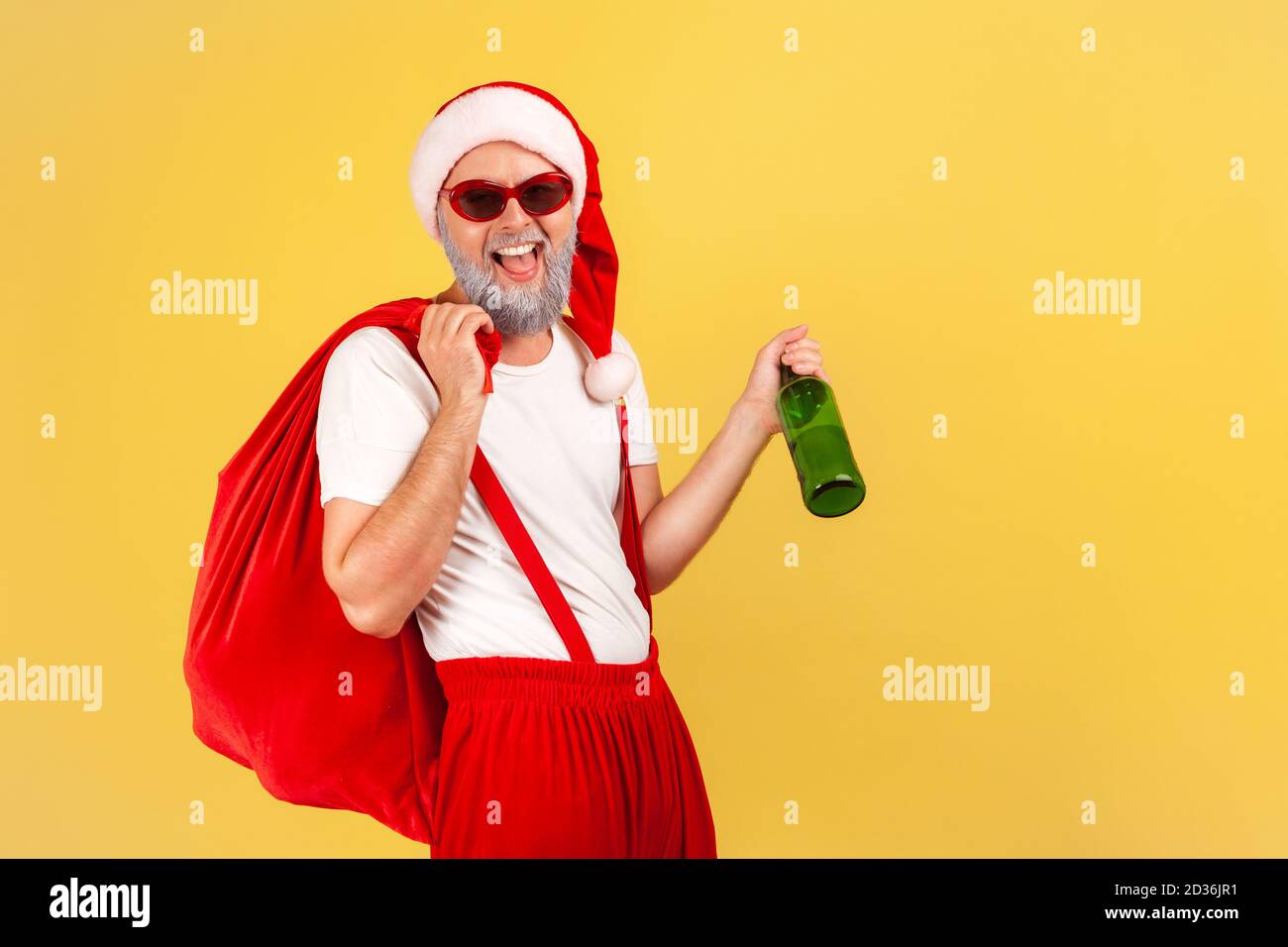 Carefree elderly man in sunglasses and santa claus costume with big red bag holding bottle with alcohol and smiling at camera, bad habits. Indoor stud Stock Photo