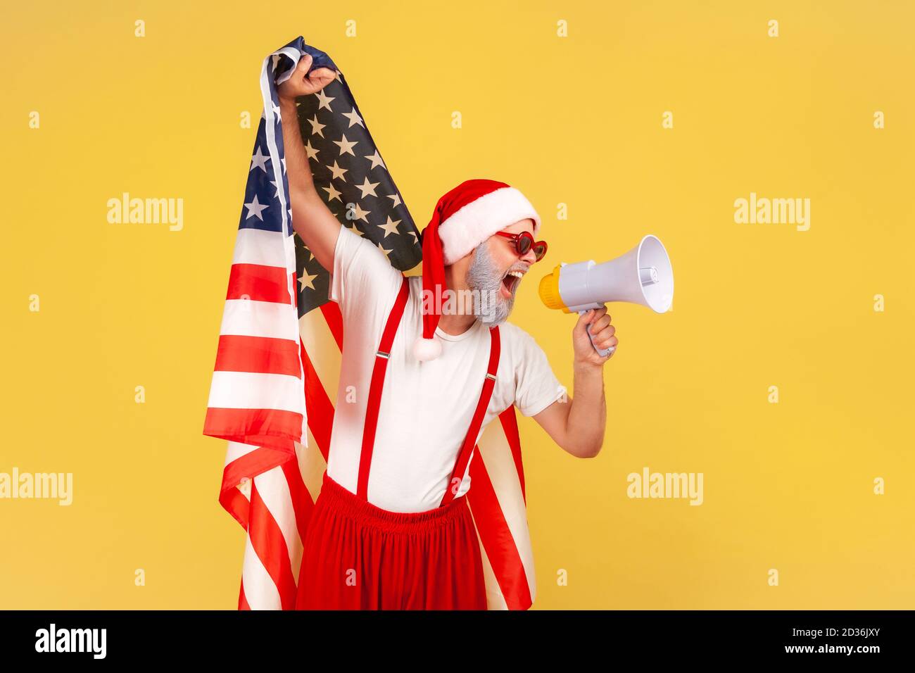 Serious elderly man in santa claus costume screaming at loudspeaker holding flag of united states of america, patriotic protest on winter holidays. In Stock Photo