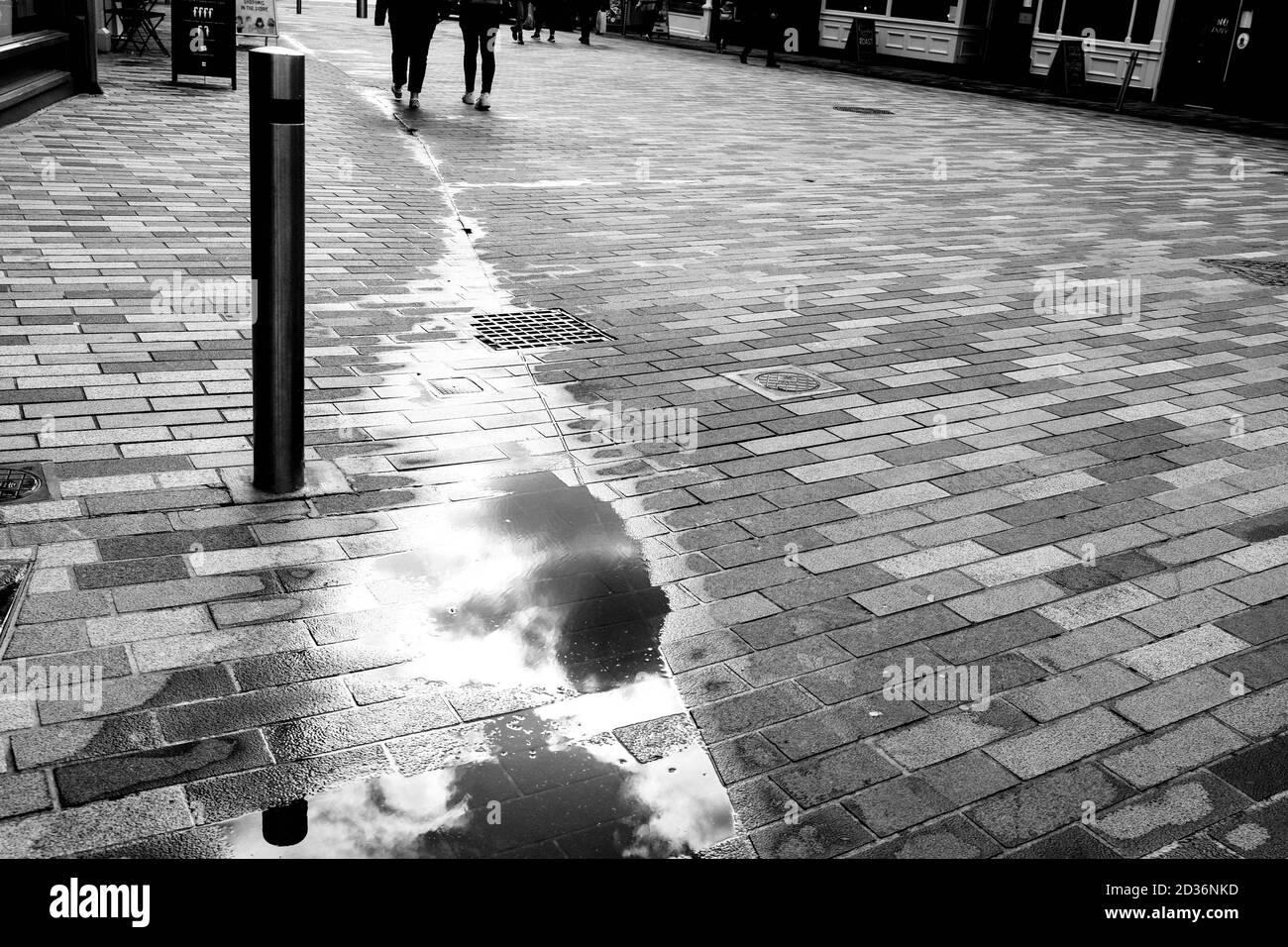 Two People Walking In The Distance On A pavement With Standing Water Puddles Reflection of Clouds in the Sky Stock Photo