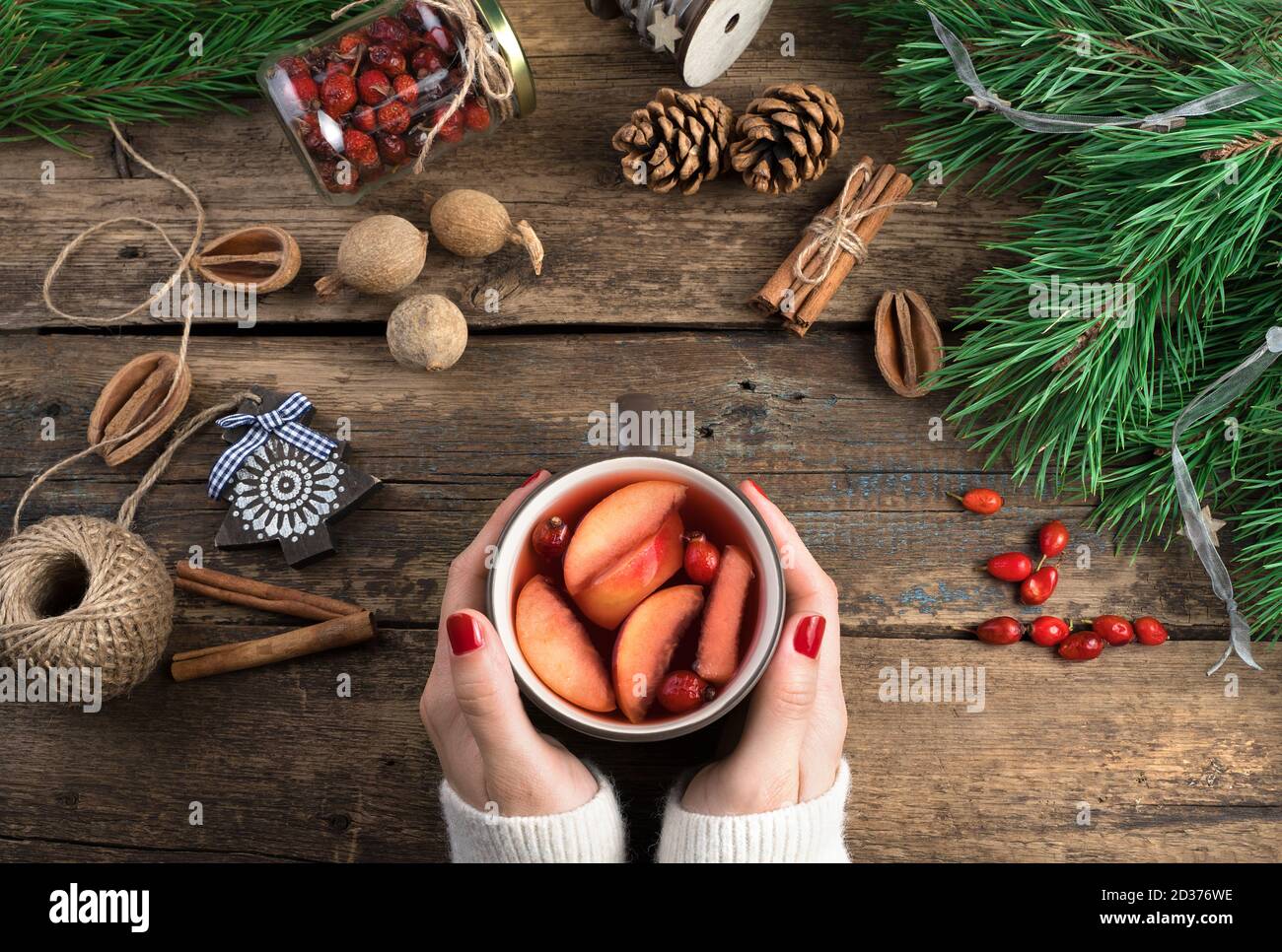 Women's hands hold a mug of mulled wine on a dark wooden background surrounded by Christmas toys, berries, cinnamon and pine branches. Stock Photo