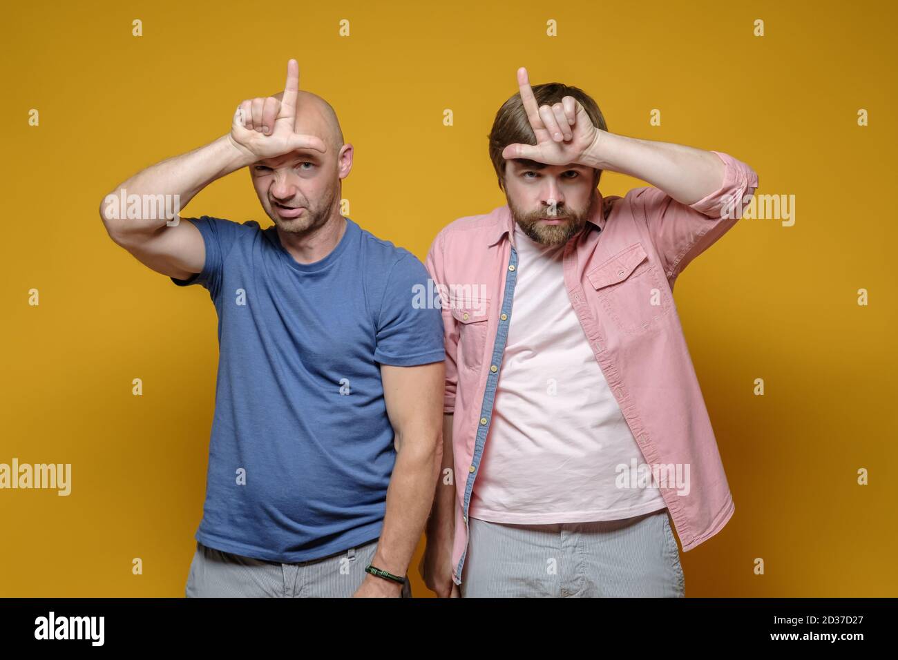 Two serious men in casual clothes are making mocking of someone by making a loser gesture with their fingers on their foreheads.  Stock Photo