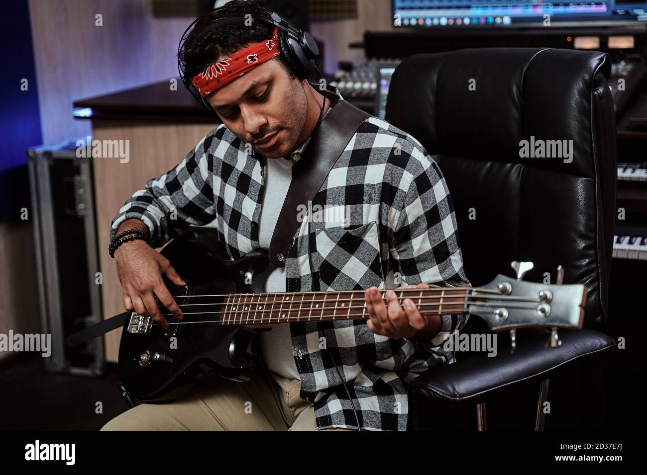 Portrait of focused young man, male artist in headphones playing guitar while sitting in recording studio Stock Photo