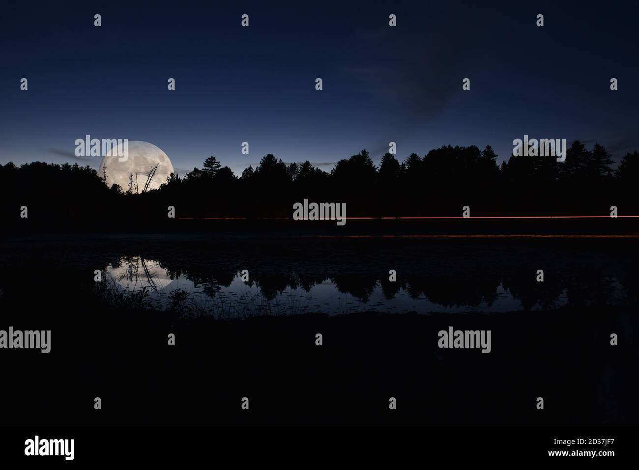 Reflection of light trails from a vehicle on a small lake at night, set against trees and night sky. Stock Photo