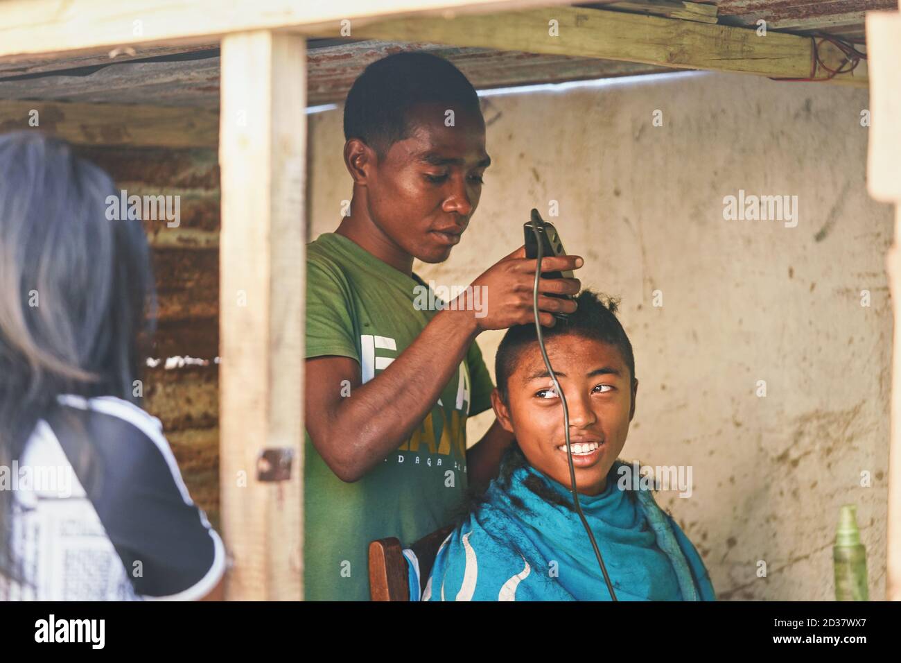 Ranohira, Madagascar - April 29, 2019: Unknown local boy getting his haircut from a friend at shop back room made from wooden boards. People in this p Stock Photo