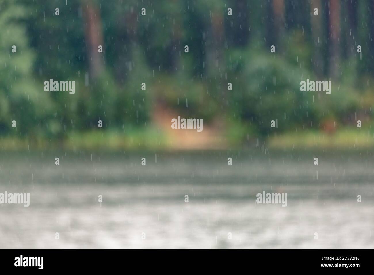 Abstract bokeh background of raindrops falling on water with a soft, blurred green forest shoreline. Stock Photo