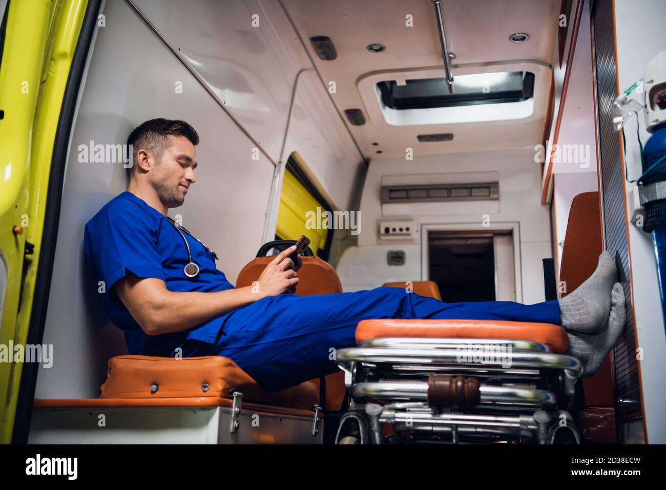 Male paramedic in a blue uniform resting and talking to someone over the video call on his smartphone in the ambulance car. Stock Photo