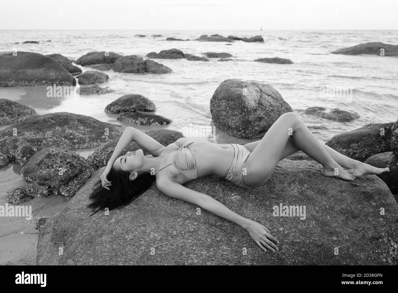 Profile view of young beautiful Asian woman lying down with eyes closed on the rocks of public beach in Hua Hin Thailand Stock Photo