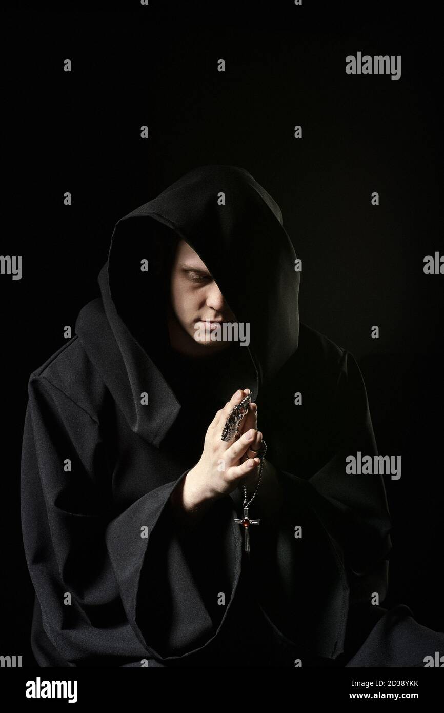 Assassin in dark clothes in church. Sorcerer in black mantle doing mysterious rite in dark room. Monk prays in darkness. Magic ritual in shadow. Sinne Stock Photo