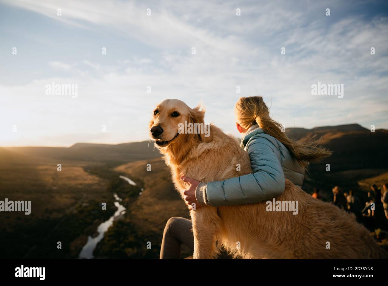 Blonde female sits on top of mountain with arm around golden puppy while looking into the distance at sunset. Stock Photo