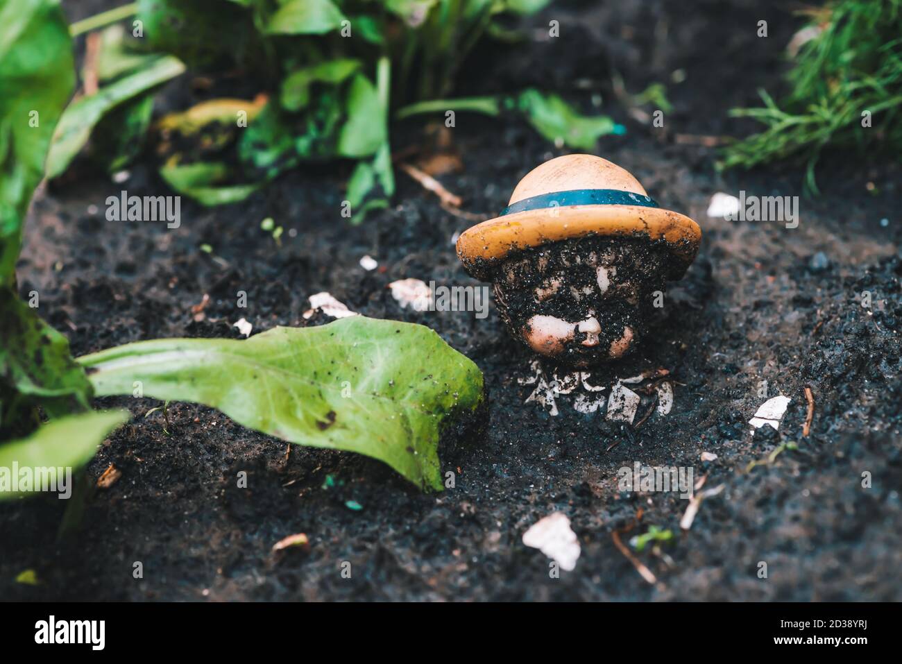 Dirty doll in soil near plants and trash. Old thrown soiled toy buried in ground. Sad pessimistic statuette in garden. Gloomy place. Melancholy backgr Stock Photo