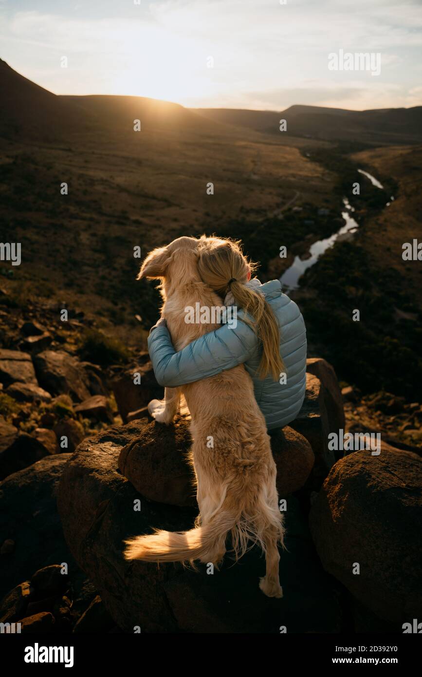 Young caucasian blonde female hugging golden dog while looking at river sitting on top of mountain. Stock Photo