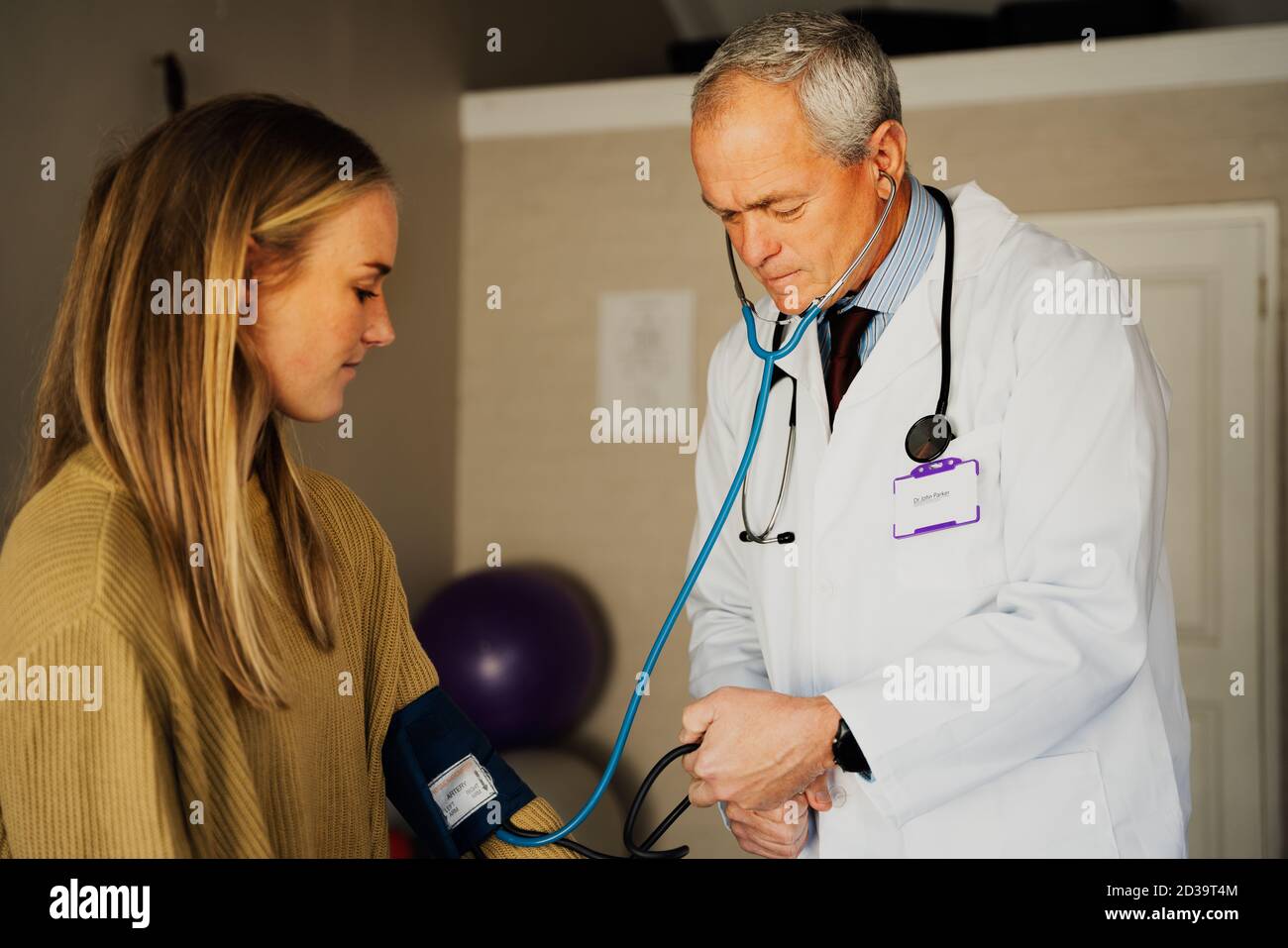Elderly male doctor gently taking blood pressure from young female patient sitting in doctors office. Stock Photo