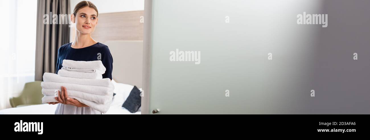 horizontal image of young maid in apron looking at camera and holding clean towels in hotel room Stock Photo
