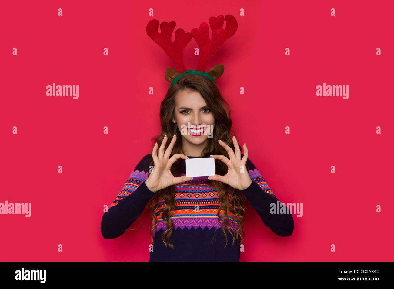 Smiling young woman in sweater with colorful pattern wearing christmas reindeer horns showing white card. Front view. Waist up studio shot on red back Stock Photo