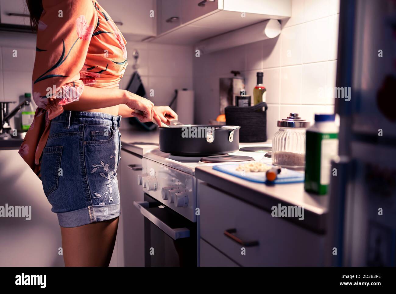 Woman cooking dinner in home kitchen at night. Healthy vegan or vegetarian diet. Stir food in frying pan on stove. Happy person preparing meal. Stock Photo