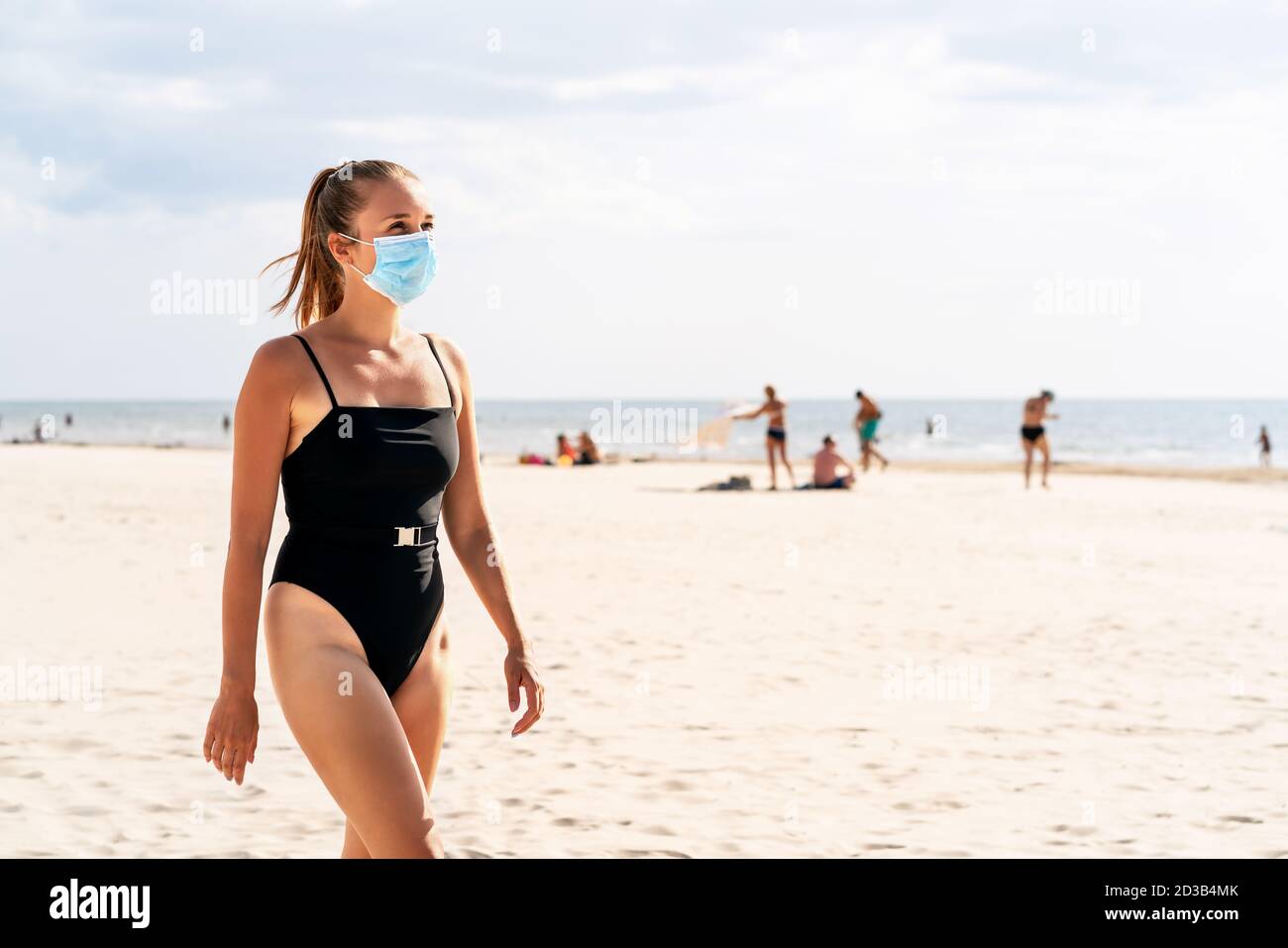 Woman walking on the beach wearing a face mask to protect from corona virus. Tourist on a vacation with people in the background. Facemask on mouth. Stock Photo