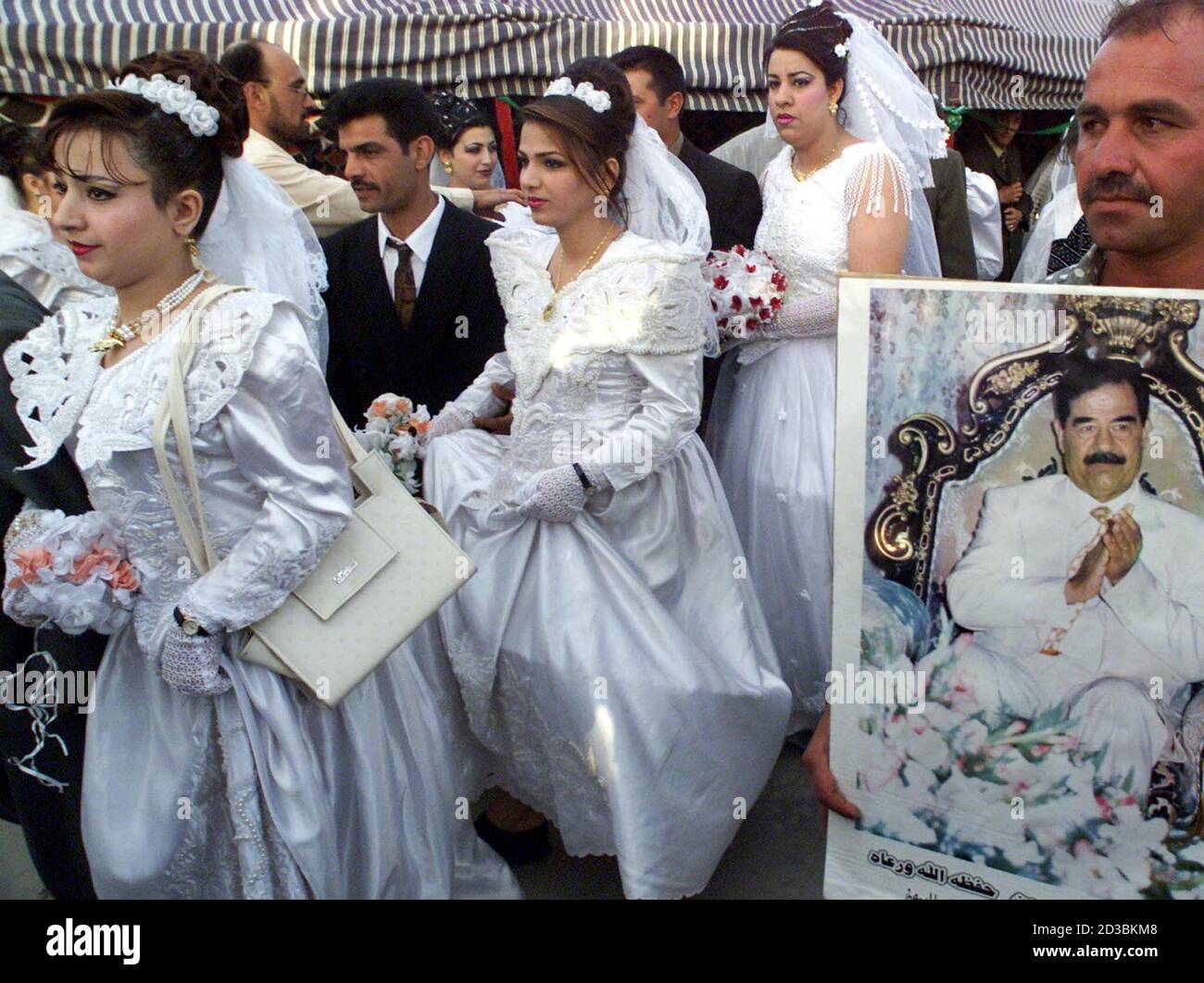 Brides and bridgegrooms pass a portrait of Iraqi President Saddam Hussein during a mass wedding of around 300 couples Baghdad, April 25, 2002 as part of the country's celebrations of the President's birthday on Sunday. Iraq's government usually arranges mass weddings to encourage Iraqis to get married.  Economic hardships because of more than 11 years of crippling sanctions make many Iraqis refrain from marriage. Stock Photo