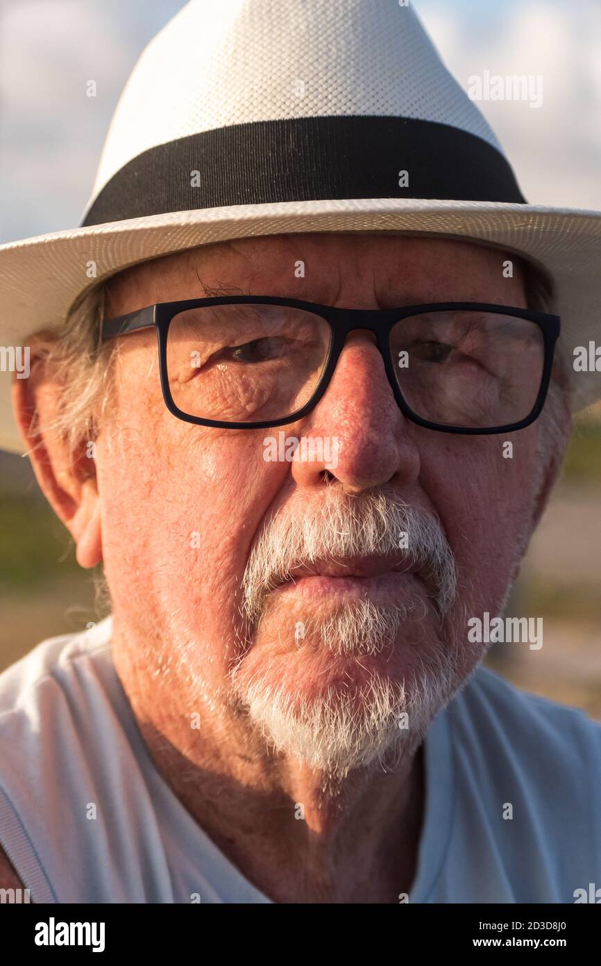 Portrait of an elderly man in a Panama hat and black-rimmed glasses whose face is half-lit by the sunset light. Stock Photo