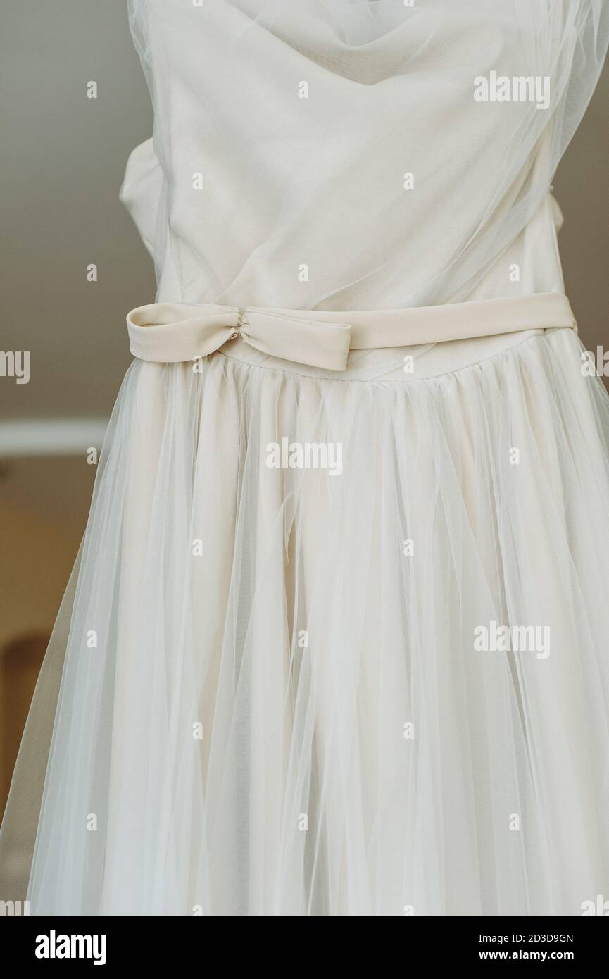 Close up of a minimalist bow on a beautiful white minimal handmade wedding dress hanging on a wooden hanger in the middle of the room. Best day of the Stock Photo