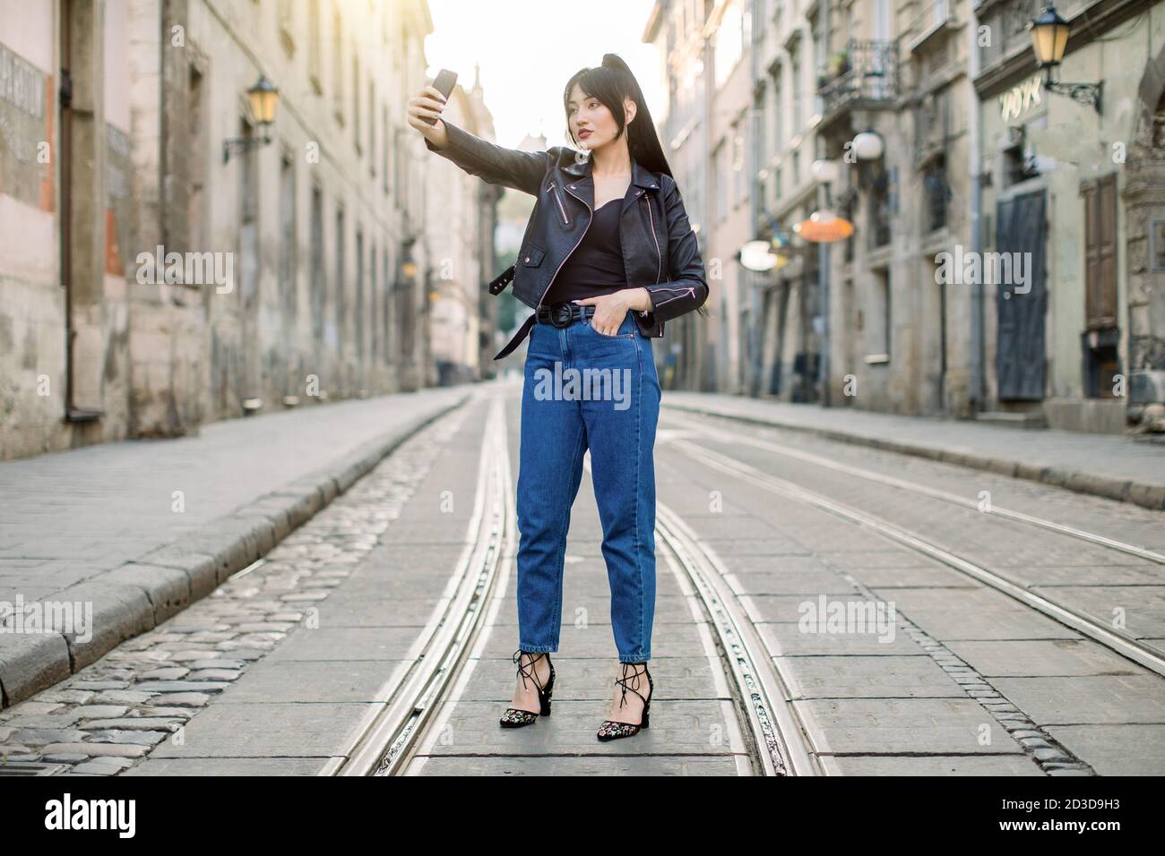 Full length horizontal shot of young cheerful Asian woman, wearing trendy denim pants and black leather jacket, taking selfie by mobile phone, posing Stock Photo