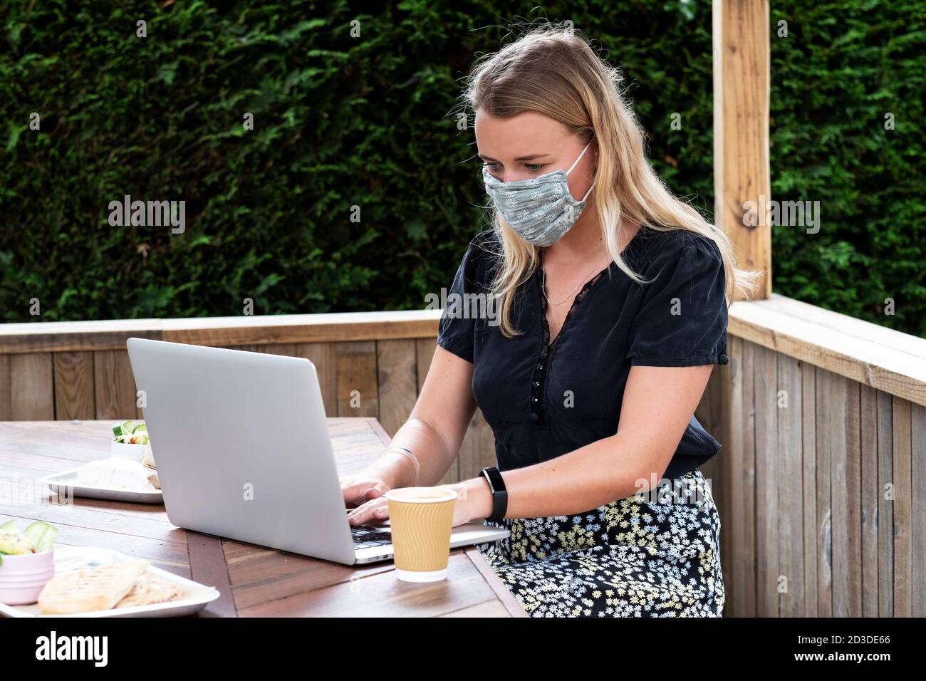 Young blond woman wearing blue face mask, sitting at table, using laptop computer. Stock Photo