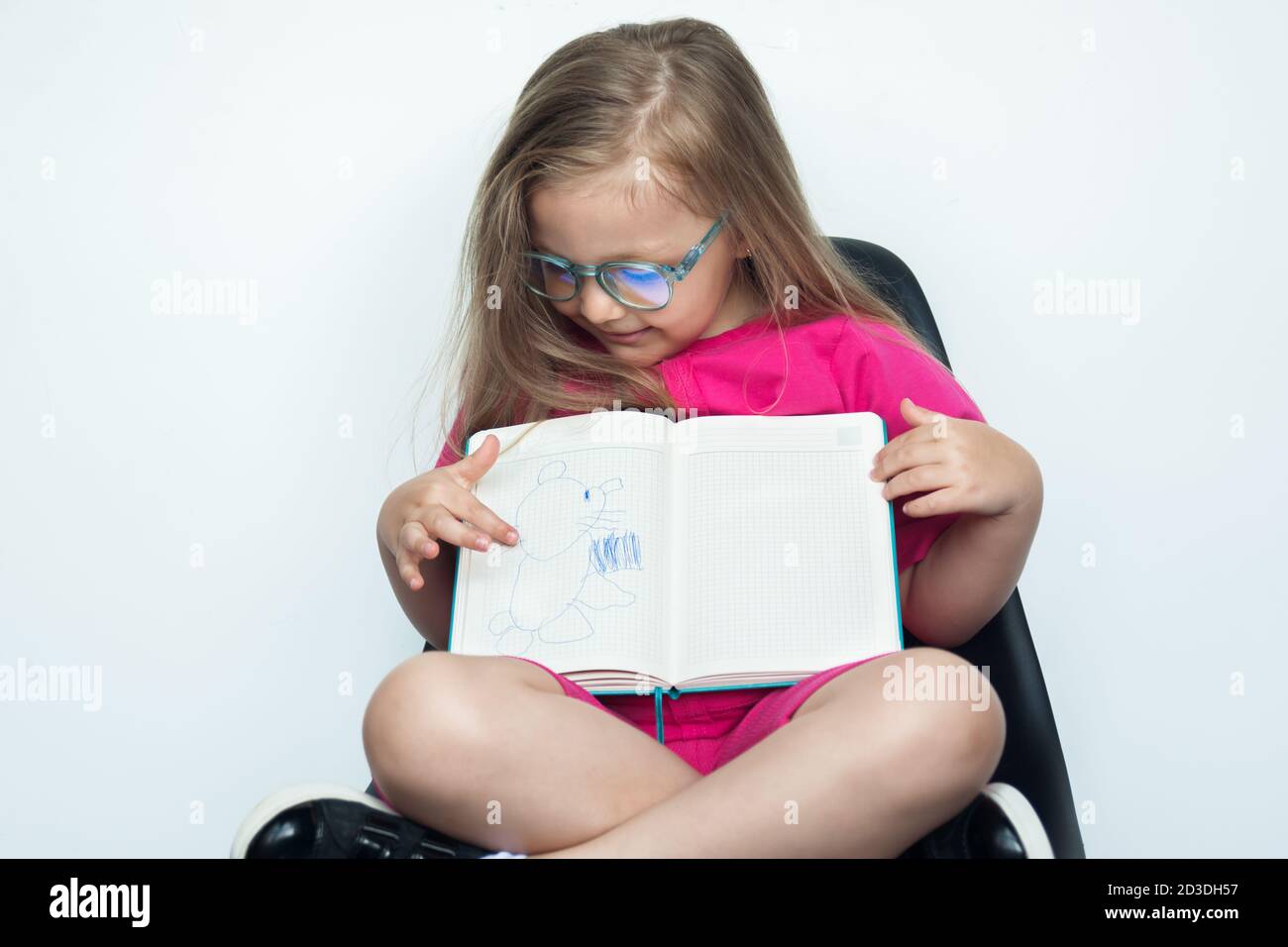 Small caucasian girl with blonde hair and glasses sitting in armchair on a white studio wall is gesturing at camera her book with a drawn picturedrown Stock Photo