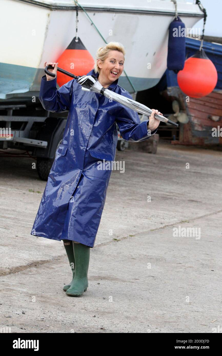 White Caucasian dyed blonde middle aged woman in her 40s wearing a blue shiny  PVC raincoat purchased from a second hand shop. Pretending to dance in her  raincoat and wellington with an