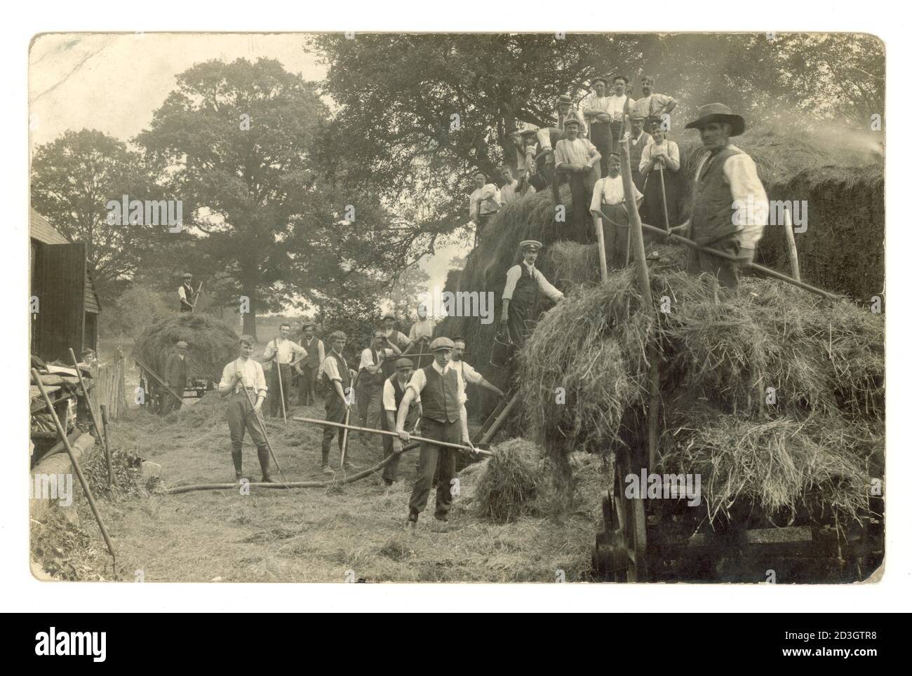 Original early 1900's, Edwardian postcard of farm labourers and fire brigade monitoring, containing and damping down a smouldering haystack, stack fire, using a hosepipe and buckets of water and pitchforks a grumpy-looking woman looks on. Circa 1906 U.K. Stock Photo