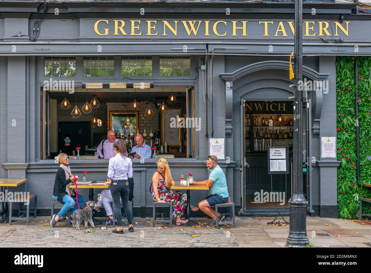 People enjoy a 'socially distanced' drink in the sunshine on the pavement outside the Greenwich Tavern near the Royal Maritime Museum in Greenwich, Lo Stock Photo