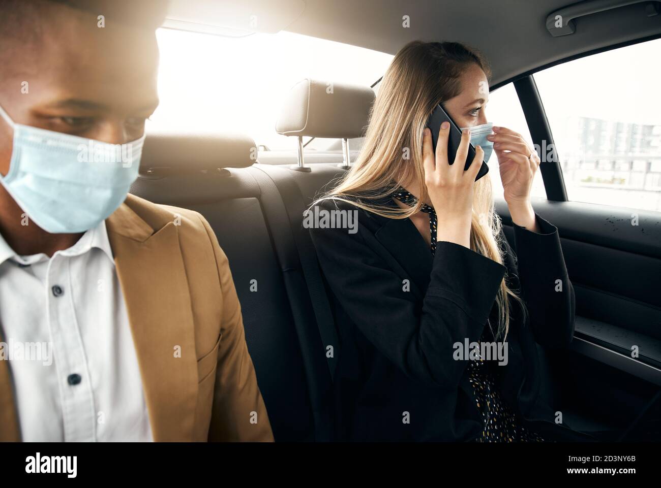 Business Couple Wearing Masks Using Laptop And Mobile Phone In Back Of Taxi During Health Pandemic Stock Photo
