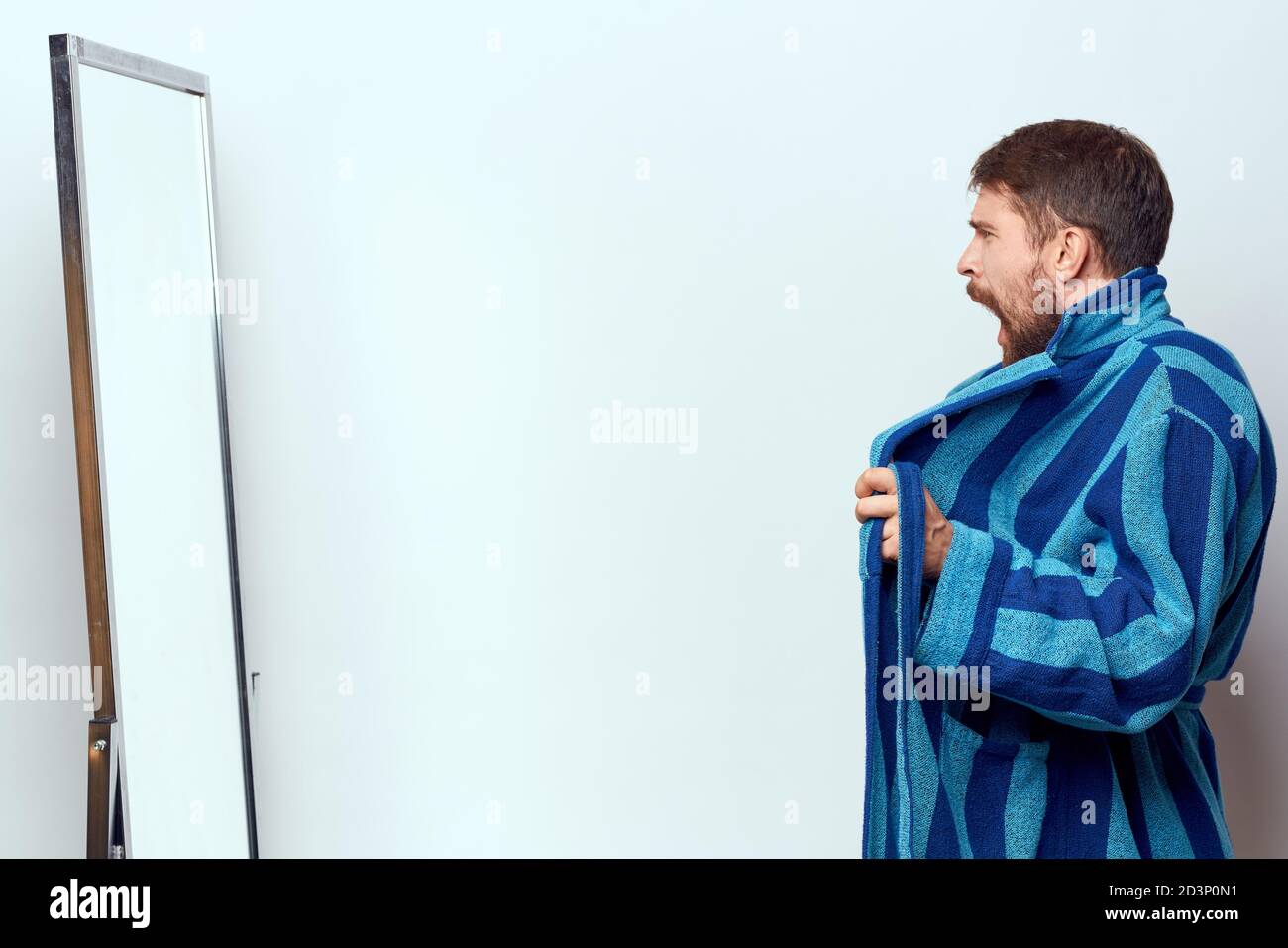 a man in a blue robe examines himself in a mirror in a bright room ...