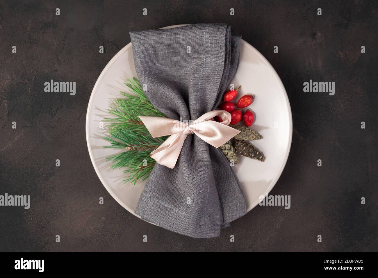 The white plate is decorated with a gray napkin with a pink bow, fir branches and a Christmas ball on a light concrete background. Stock Photo