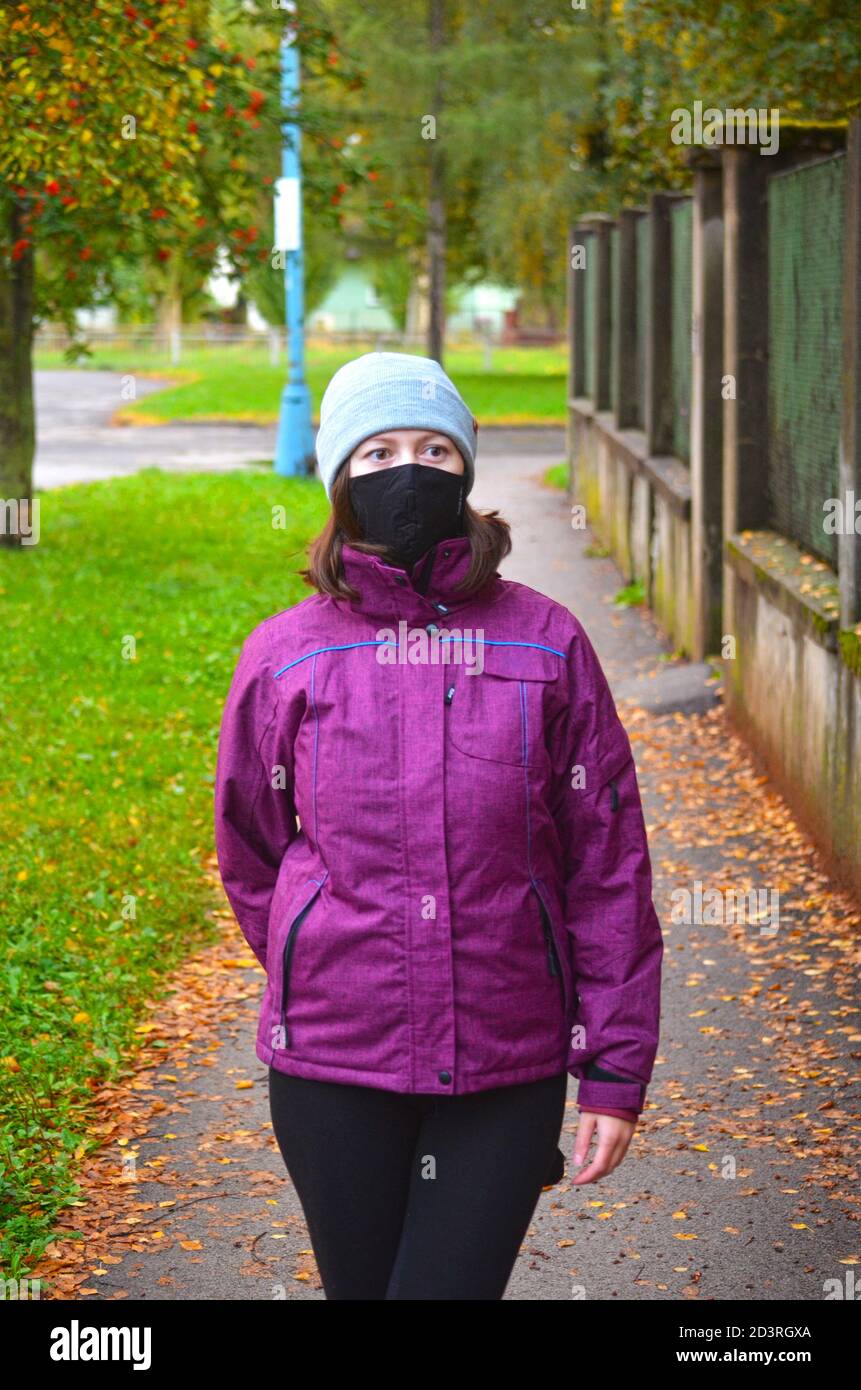 Young woman in warm clothes wearing purple jacket with black reusable face mask and knit hat standing on sidewalk covered with brown leaves. Autumn Stock Photo