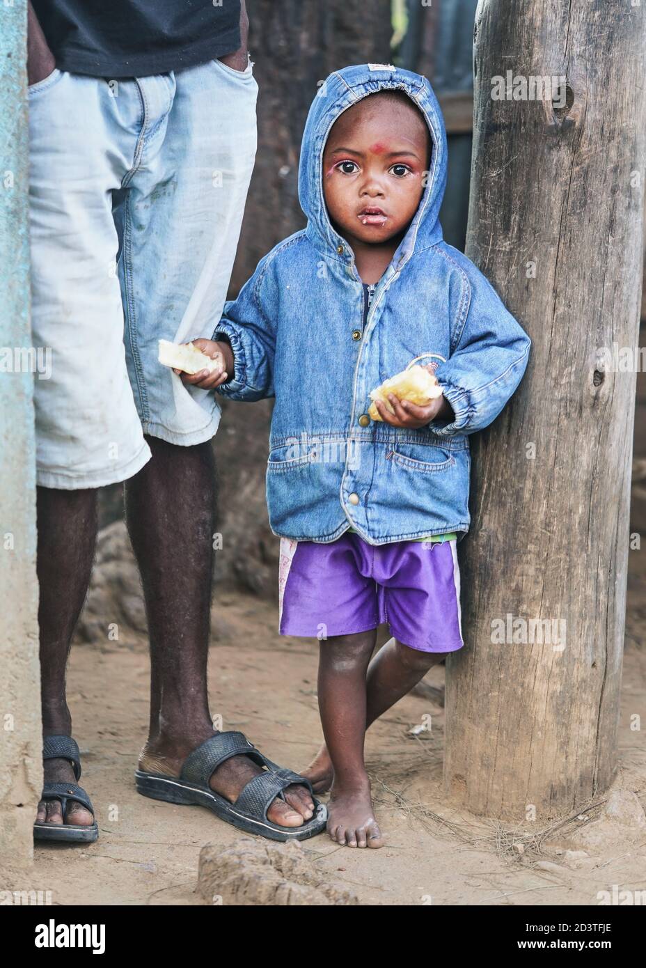 Ranohira, Madagascar - April 29, 2019: Unknown Malagasy boy, in blue denim jacket with hood and shorts, barefoot, standing next to his father legs, ho Stock Photo