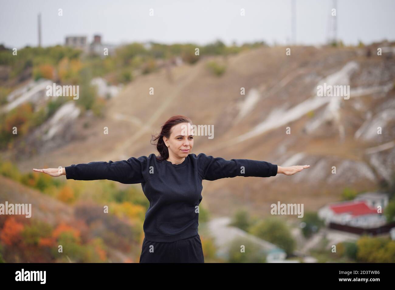Young woman doing warm-up in hilly terrain. Adult female in black casual clothes doing sports in fresh air in countryside. Stock Photo