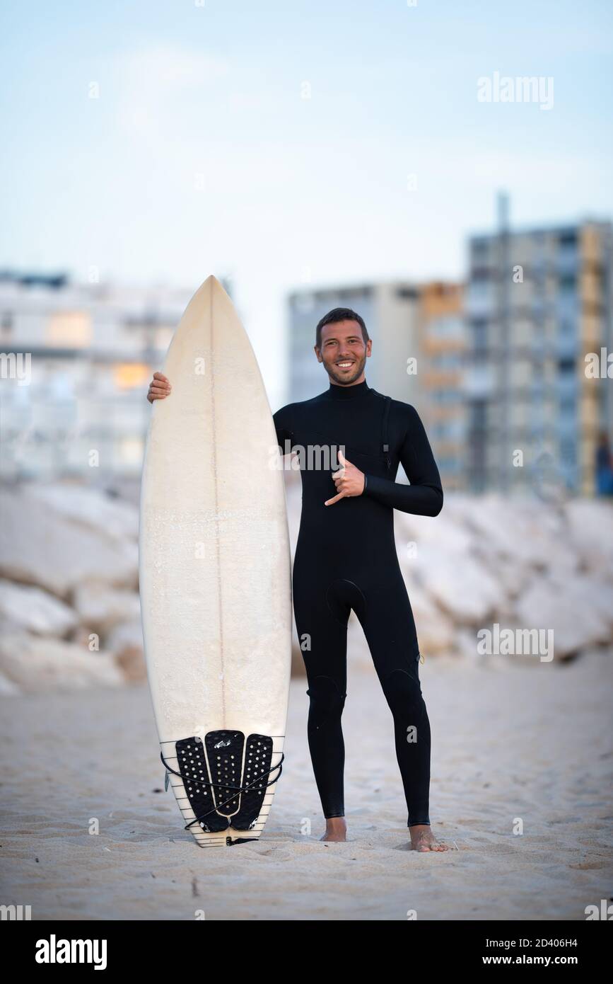 Young smiling surfer in black zipless surfing wetsuit with shortboard showing shaka sign in