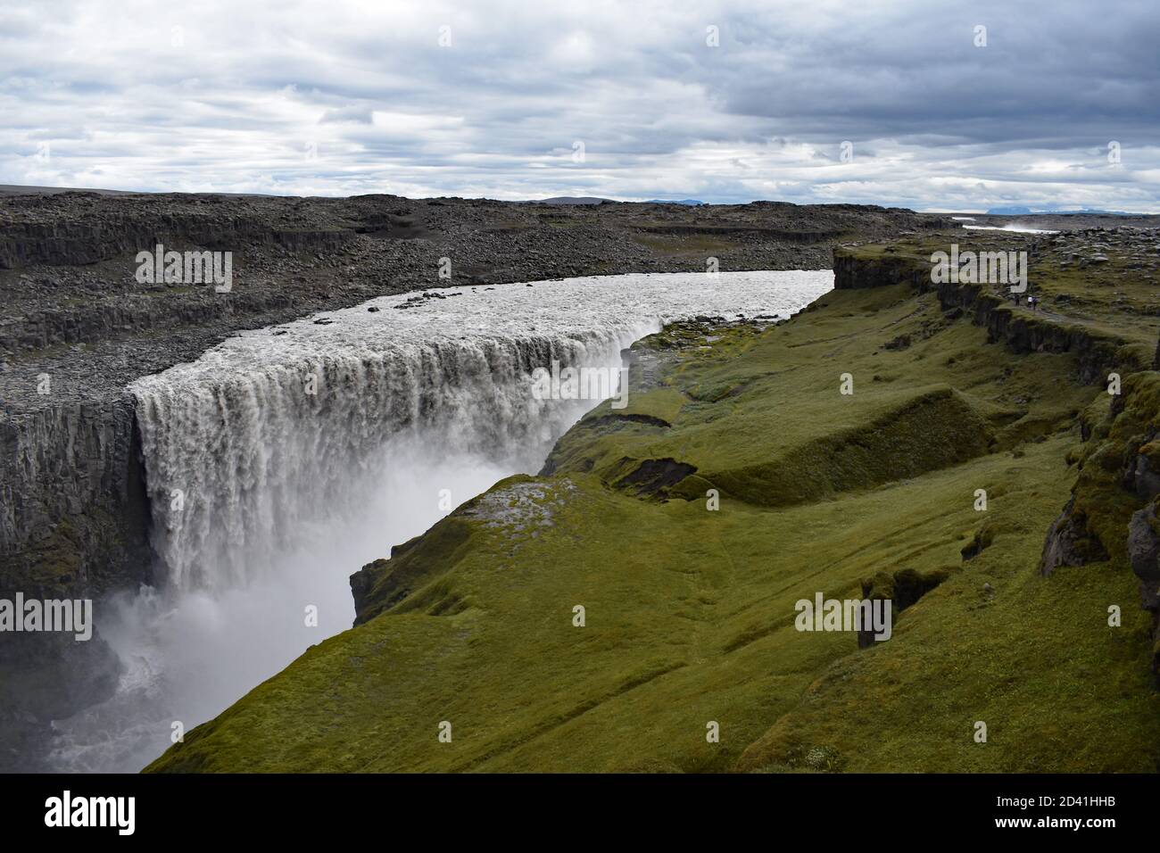 A view of Dettifoss Waterfall in Northeast Iceland. The green eastern side of the canyon contrasts grey and barren western side and rushing grey water. Stock Photo