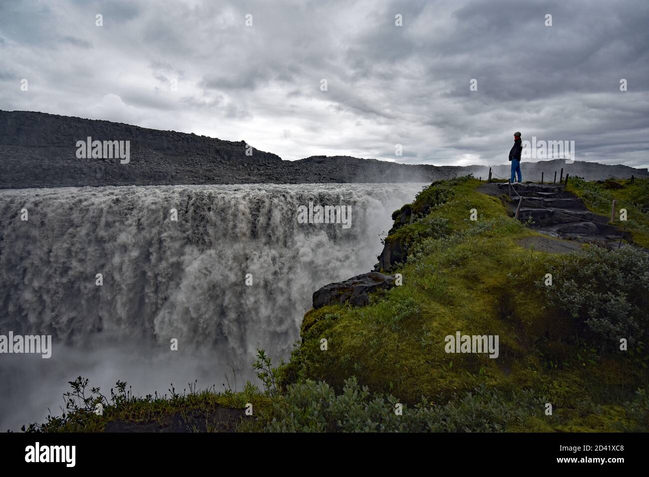 A male wearing a hooded coat standing on stone steps near the edge of Dettifoss Waterfall in Northeast Iceland as it plunges into the canyon below. Stock Photo