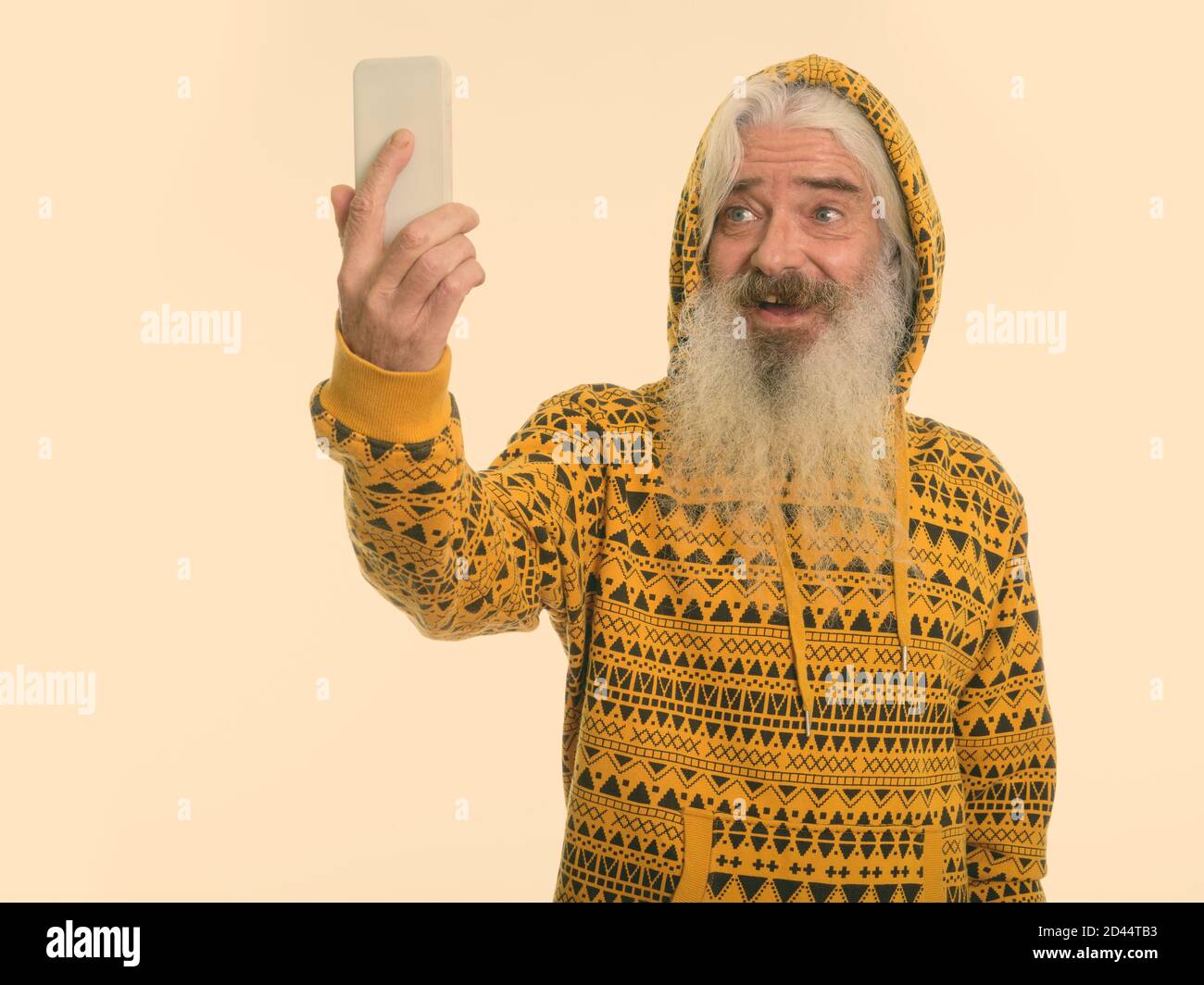 Studio shot of happy senior bearded man smiling while wearing hoodie and taking selfie picture with mobile phone Stock Photo