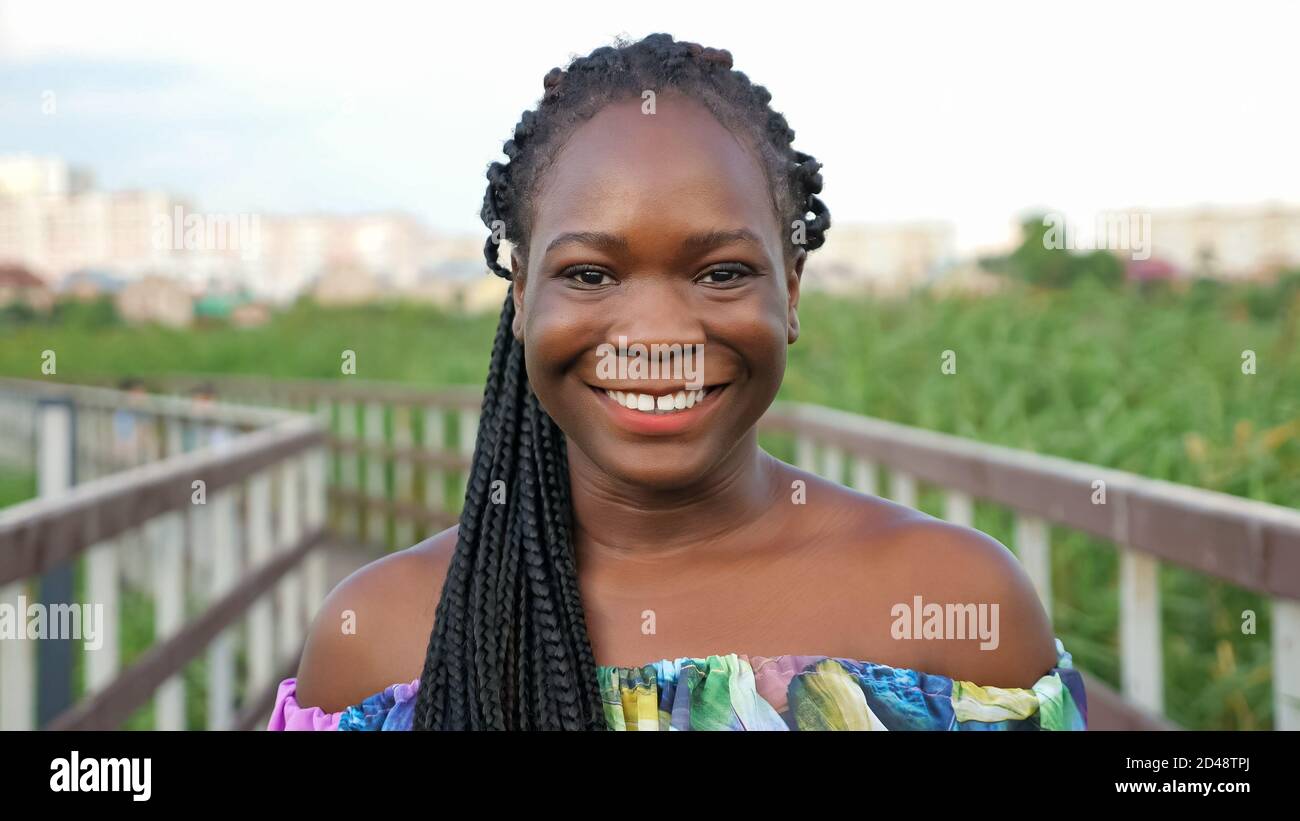 attractive African-American girl with long cornrows in bright color blouse smiles standing on wooden bridge in green reeds closeup Stock Photo