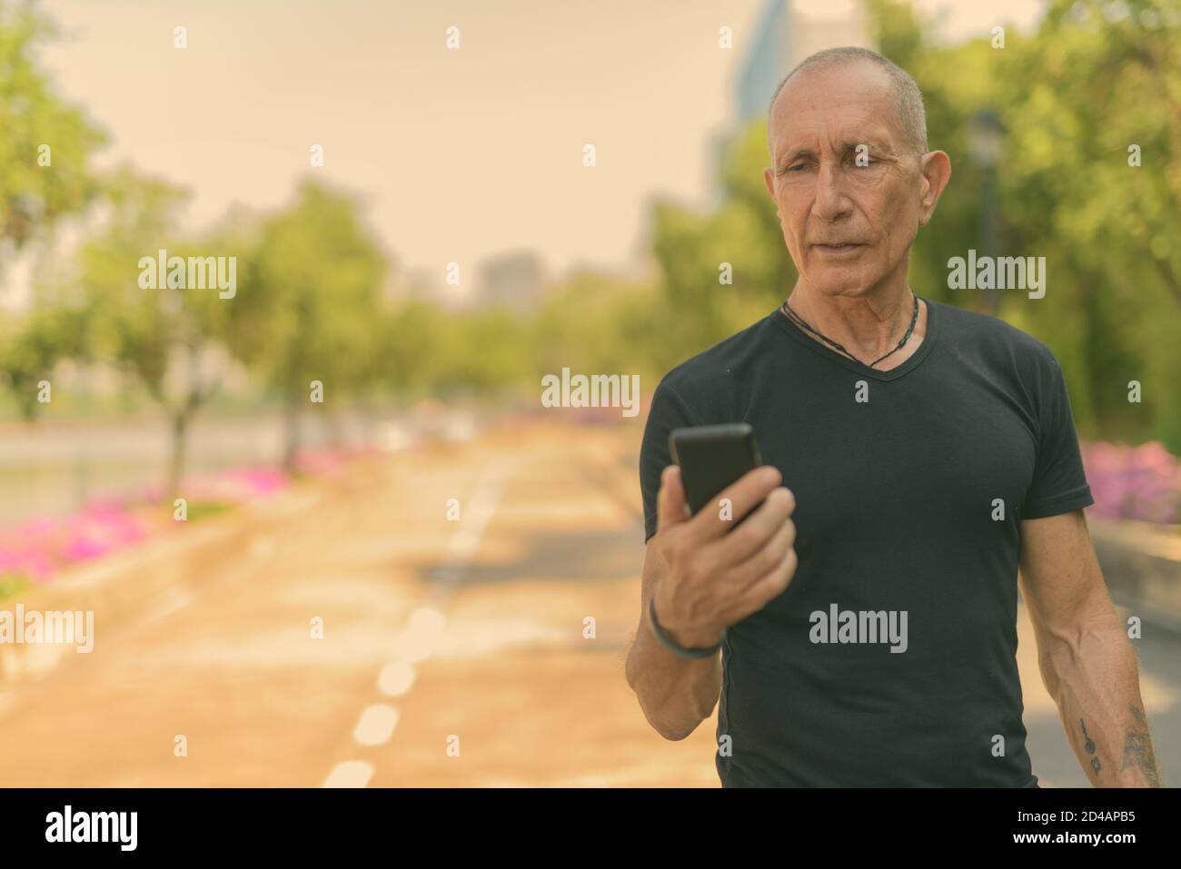 Bald senior tourist man using mobile phone on the side of the street at peaceful park in Bangkok Thailand Stock Photo
