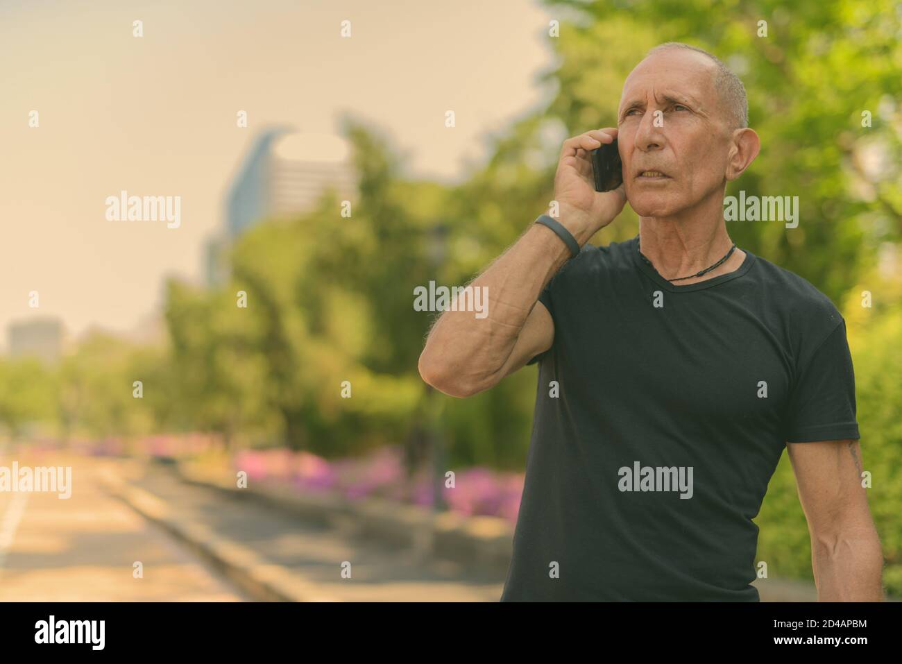 Bald senior tourist man thinking while talking on mobile phone against view of the street at peaceful park in Bangkok Thailand Stock Photo