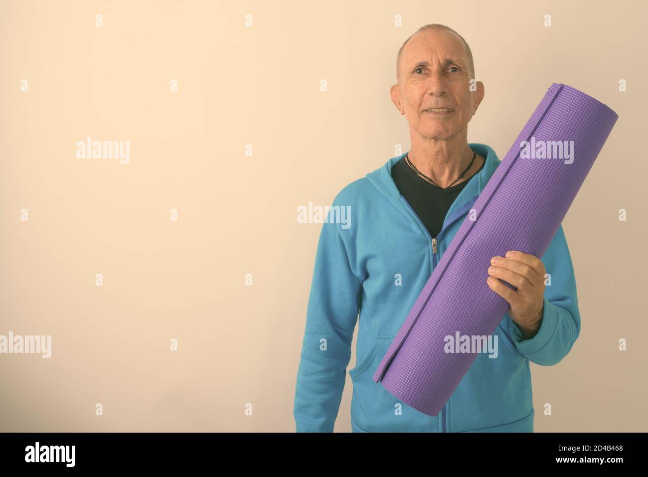 Studio shot of happy bald senior man smiling while holding yoga mat ready for gym against white background Stock Photo