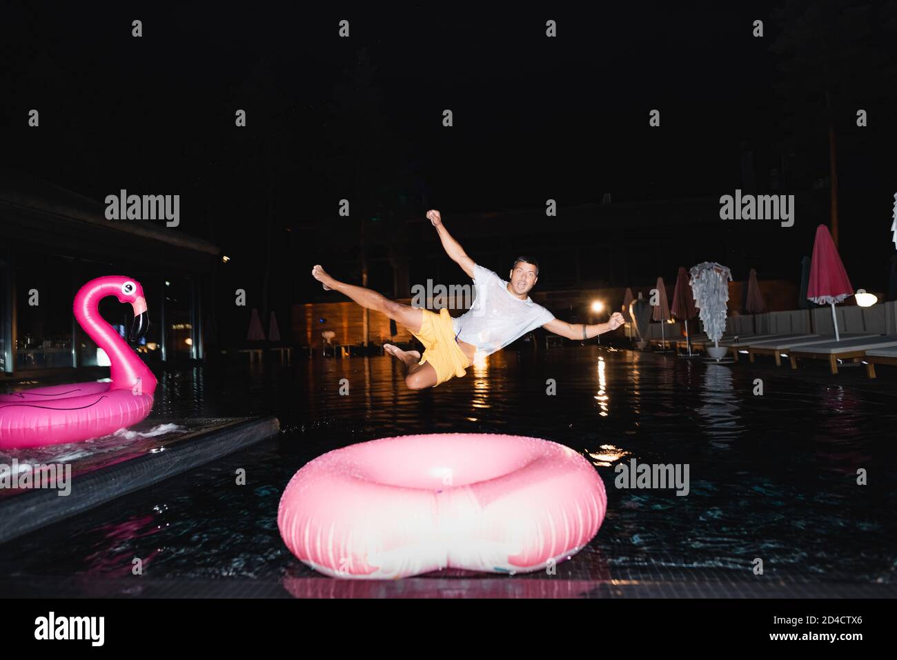 Selective focus of young man looking at camera while jumping in swimming pool at night Stock Photo